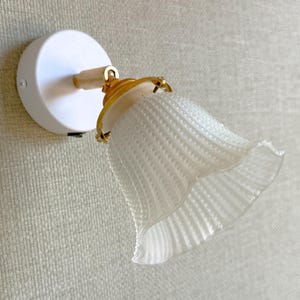 May include: A wall-mounted light fixture featuring a circular white base, a gold-toned adjustable joint, and a translucent, fluted glass shade with a scalloped edge. The lamp includes a small black power switch located on the side of the white base.