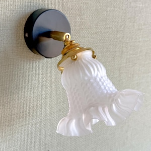 May include: A wall-mounted light fixture featuring a circular black base, a brass-toned metal arm, and a frosted glass shade. The shade has a textured, scale-like pattern on the upper portion and a ruffled, flared edge at the bottom opening.