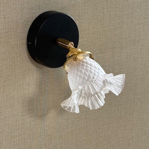 May include: A wall-mounted light fixture with a black circular base and a brass-colored arm. The light has a white, textured glass shade with a ruffled edge. The fixture is mounted on a textured, neutral-toned wall.