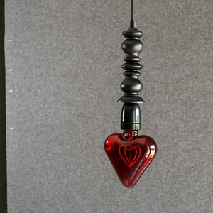 May include: A red heart-shaped light bulb hangs from a black cord and a black metal fixture. The bulb has a filament shaped like a heart.