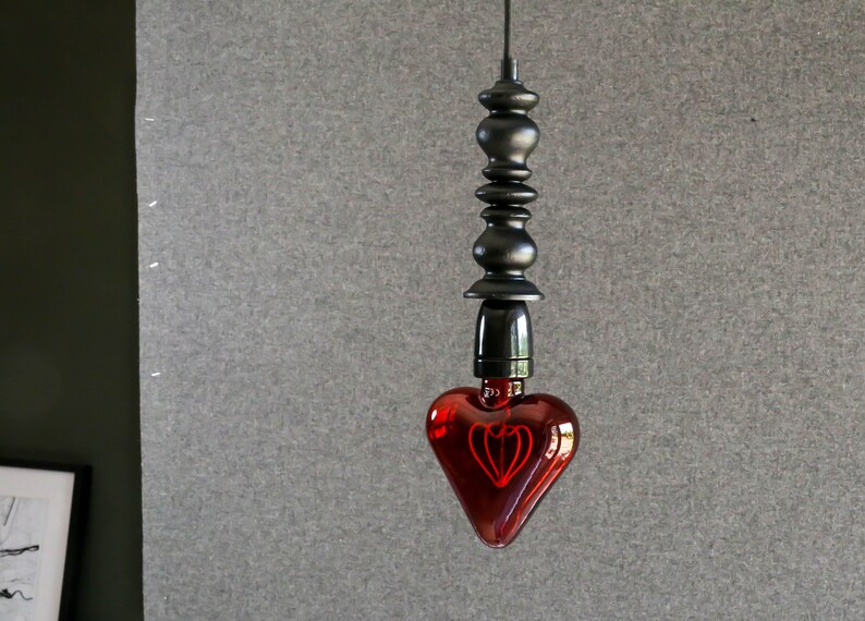 May include: A red heart-shaped light bulb hangs from a black cord and a black metal fixture. The bulb has a filament shaped like a heart.