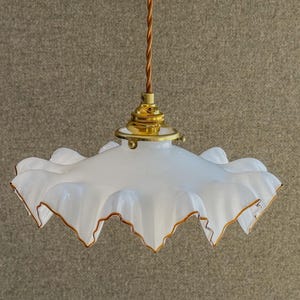 Könnte beinhalten: Eine Pendelleuchte mit einem weißen, gerüschten Glasschirm, der an den gewellten Rändern mit einem zarten goldfarbenen Rand versehen ist. Der Schirm ist an einer messingfarbenen Metallfassung befestigt und hängt an einem gedrehten, braunen stoffummantelten Kabel.