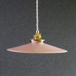 Peut inclure: Suspension luminaire avec un abat-jour rose en forme de soucoupe. Le luminaire a une base couleur laiton et un cordon torsadé blanc. La lumière est suspendue sur un fond gris.
