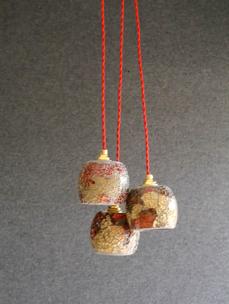 May include: Three glass pendant lights with red and gold mosaic patterns hang from red cords against a gray background.