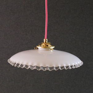 Peut inclure: Suspension luminaire en verre blanc avec bord festonné. Le luminaire a une base dorée et un cordon rose. La lumière est suspendue sur un fond gris foncé. Le design général est vintage et décoratif.