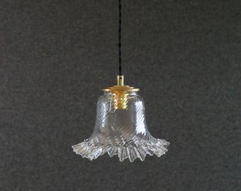 Op de afbeelding: Transparante glazen hanglamp met een geschulpte rand en goudkleurige hardware. De lamp hangt aan een zwart snoer tegen een donkergrijze achtergrond. De glazen kap heeft een getextureerd, wervelend patroon.