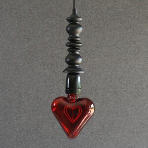 May include: A red heart-shaped glass light bulb hangs from a black metal cord and fixture. The bulb has a decorative filament inside that creates a heart shape when lit.