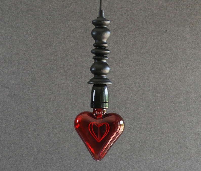 May include: A red heart-shaped glass light bulb hangs from a black metal cord and fixture. The bulb has a decorative filament inside that creates a heart shape when lit.