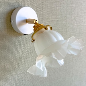 May include: A wall-mounted light fixture featuring a circular white base, a gold-toned metal arm, and a frosted glass shade. The shade has a fluted, petal-like design with ruffled edges, attached to the arm by a gold-toned metal hardware fitting.