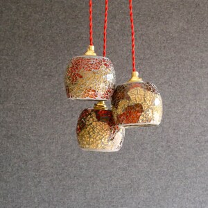 May include: Three hanging pendant lights with red cords. The lights are made of glass with a mosaic pattern in shades of gold and red.