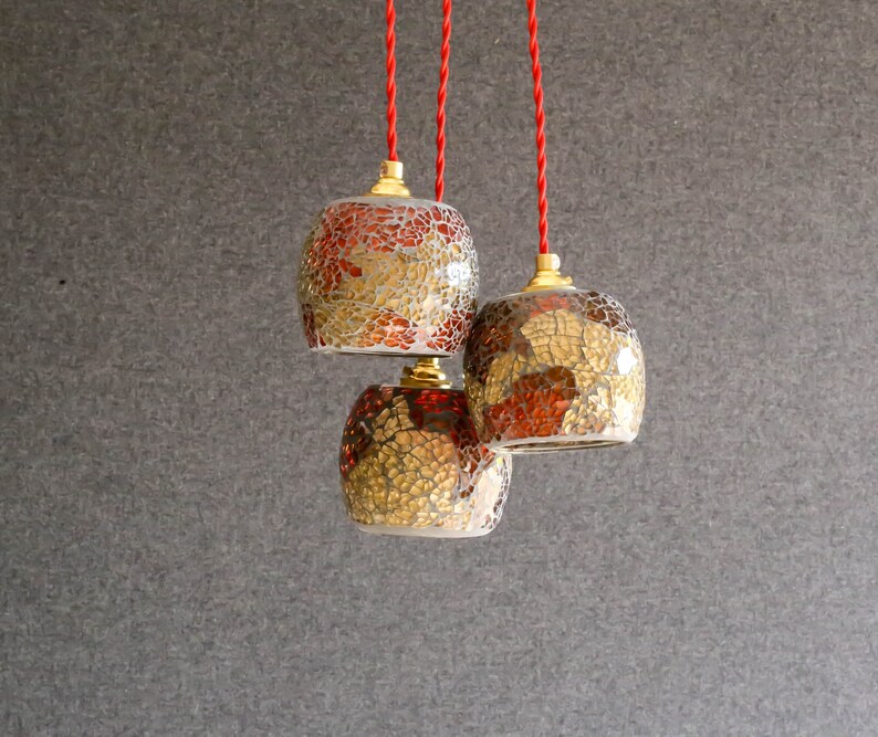May include: Three hanging pendant lights with red cords. The lights are made of glass with a mosaic pattern in shades of gold and red.
