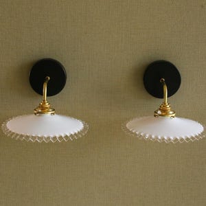 Puede incluir: Dos apliques de pared a juego. Cada aplique presenta una base circular negra, un brazo dorado curvado y una pantalla blanca con borde festoneado. Los apliques están montados en una pared verde oliva texturizada.