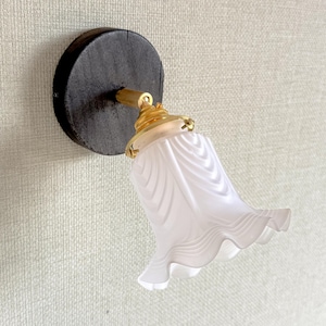 May include: A wall-mounted light fixture featuring a circular dark wood base, a metallic gold-toned arm, and a bell-shaped frosted glass shade with a ruffled, wavy edge. The lamp is attached to a textured, light-colored wall surface.