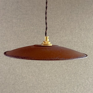Peut inclure: Une suspension luminaire dotée d'un abat-jour large, plat et circulaire avec une finition texturée brun rouille foncé. L'abat-jour est fixé à une douille en métal couleur laiton et suspendu par un fin cordon électrique torsadé marron.