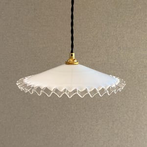 Peut inclure: Une suspension blanche dotée d'un abat-jour conique large et peu profond avec un bord en verre transparent ondulé et translucide. L'abat-jour est fixé à une monture en métal doré et suspendu par un cordon électrique noir torsadé en tissu.