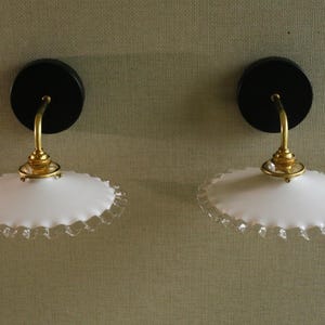 Puede incluir: Dos apliques de pared con bases circulares negras, brazos de latón curvados y pantallas de vidrio blanco con borde festoneado. Montados en una pared texturizada, ofrecen una solución de iluminación clásica.