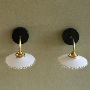 Puede incluir: Dos apliques de pared vintage. Cada uno presenta una base circular negra, un brazo curvo de latón y una pantalla de vidrio blanco con borde festoneado. Los apliques están montados en una pared texturizada de color verde claro.
