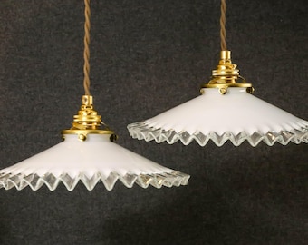 May include: Two pendant lights featuring white, conical glass shades with ruffled, clear glass edges. Each shade is attached to a polished brass fixture and hangs from a brown, twisted fabric cord against a dark, textured background.
