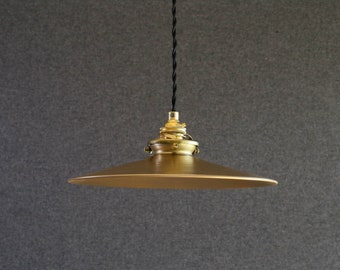 Puede incluir: Una lámpara colgante de metal de color dorado con un cable negro que cuelga del techo. La luz tiene una pantalla ancha, plana y circular.