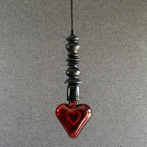 May include: A red heart-shaped light bulb hangs from a black cord and a black metal fixture. The bulb is lit and glows red.