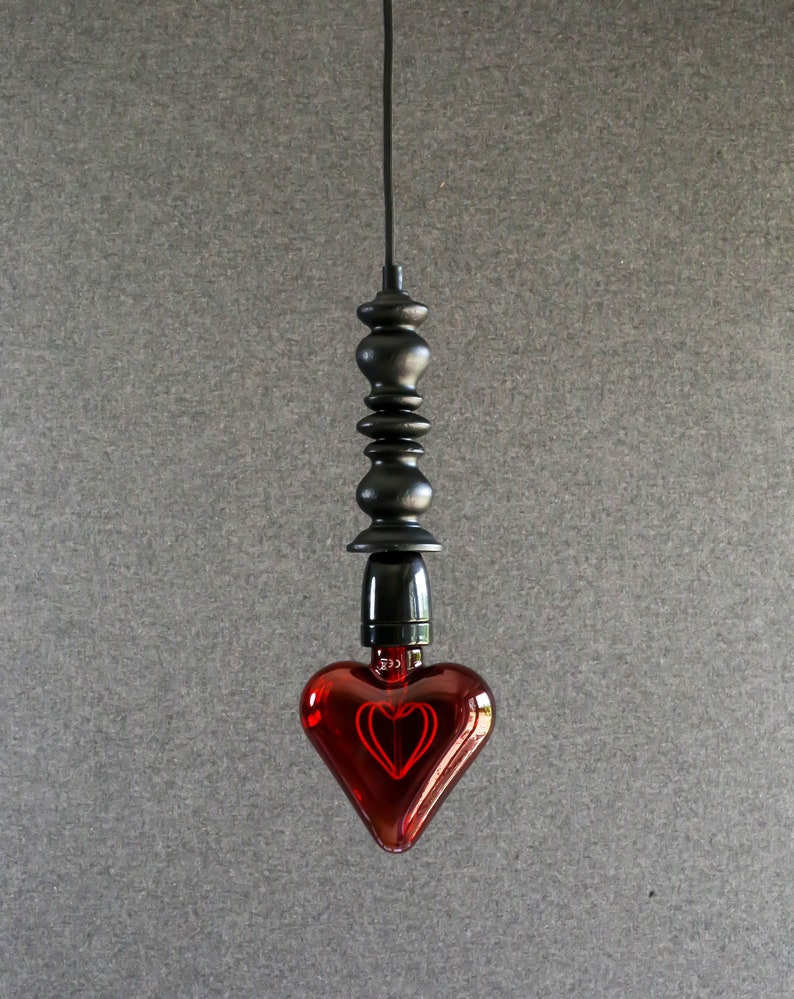 May include: A red heart-shaped light bulb hangs from a black cord and a black metal fixture. The bulb is lit and glows red.