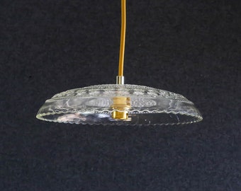Op de afbeelding: Transparante glazen hanglamp met een geschulpte rand en decoratieve details. De lamp hangt aan een gele draad en heeft een goudkleurige fitting. De achtergrond is donkergrijs.