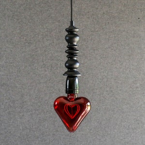 May include: A red heart-shaped light bulb hangs from a black cord and a black metal fixture. The bulb is lit and glows red.