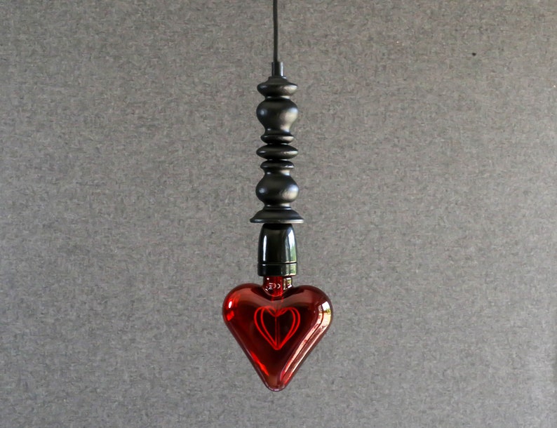 May include: A red heart-shaped light bulb hangs from a black cord and a black metal fixture. The bulb is lit and glows red.