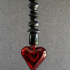 May include: A red heart-shaped light bulb hangs from a black cord and a black wooden pendant. The bulb has a red filament in the shape of a heart.