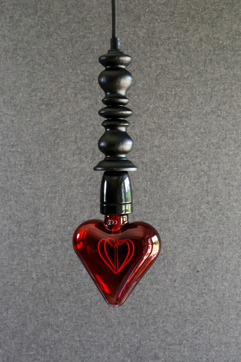 May include: A red heart-shaped light bulb hangs from a black cord and a black wooden pendant. The bulb has a red filament in the shape of a heart.
