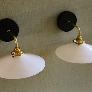 Puede incluir: Dos apliques de pared con base circular negra, brazo curvo de latón y pantalla de cristal blanco ovalada. Montados sobre una pared texturizada de color verde claro.