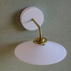 May include: A white wall light with a brass arm and a white, oval-shaped shade. The light has a circular base and a curved arm. The fixture is designed for indoor use and is mounted on a wall.