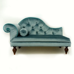 May include: A teal velvet chaise longue with a rolled arm and button tufting. The chaise features a bolster pillow and is trimmed with teal rope. The legs are dark wood.