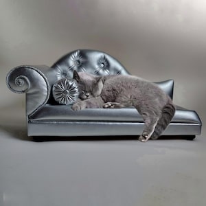 May include: A gray cat sleeping on a small, silver-colored pet sofa. The sofa has a tufted back, a decorative rolled arm, and a small, round pillow. The cat is curled up on the sofa, appearing relaxed and comfortable.