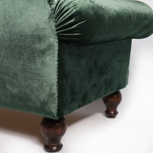 May include: A dark green velvet upholstered footstool featuring a rolled armrest with gathered fabric detailing. The piece is supported by four dark brown turned wooden legs with a rounded, bulbous shape at the base and a smooth finish.