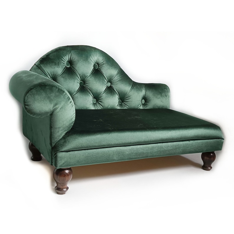 May include: A dark green velvet chaise longue featuring a tufted backrest with button detailing, a single rolled armrest on the left side, and four dark brown wooden feet. The piece has a smooth seat cushion and a curved back design.