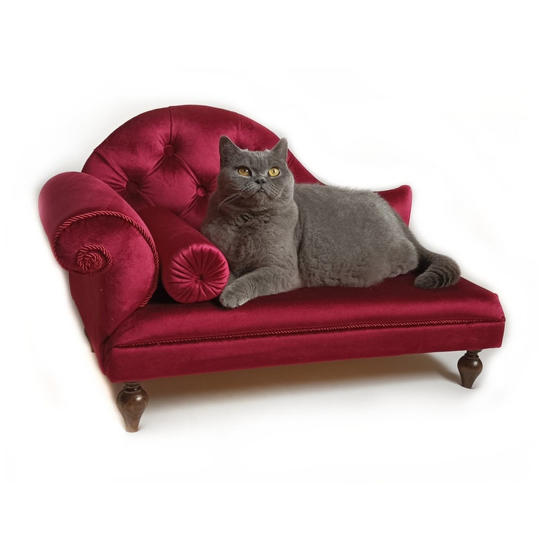 May include: A grey cat rests on a miniature red velvet chaise longue. The chaise longue has a buttoned back, rolled armrests, and a bolster cushion. The cat has yellow eyes and is looking towards the viewer. The chaise longue has wooden legs.