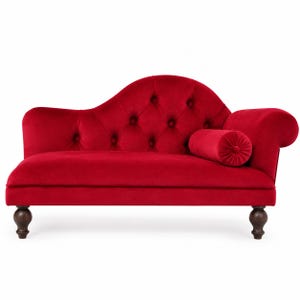 May include: A bright red velvet chaise longue featuring a tufted backrest with button detailing, a curved silhouette, a matching cylindrical bolster pillow, and four dark brown wooden legs with a rounded, turned design at the base.