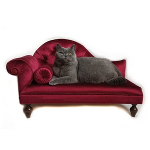 May include: A grey cat reclines on a miniature, deep red velvet chaise longue. The chaise features a buttoned back, a scrolled arm, and a small bolster cushion. The cat has yellow eyes and is looking directly at the viewer.