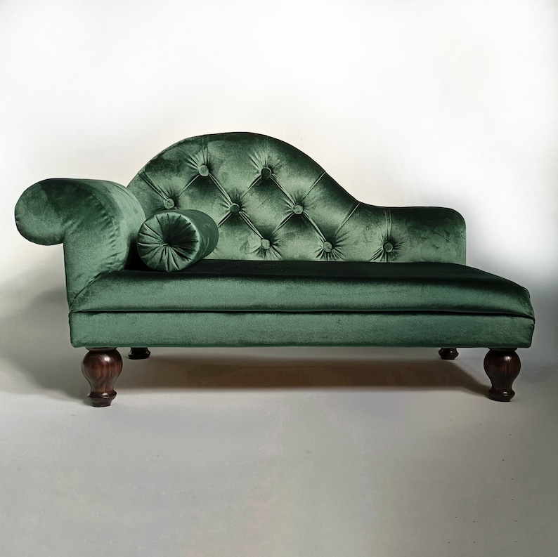 May include: A dark green velvet chaise longue featuring a tufted backrest with button detailing, a curved armrest, and a matching cylindrical bolster pillow. The furniture piece is supported by four dark brown turned wooden legs.