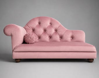 May include: A pink velvet chaise longue featuring a tufted backrest with button detailing, a curved armrest, and a matching round bolster cushion. The furniture piece is supported by four dark brown wooden feet and has a smooth, upholstered seat.