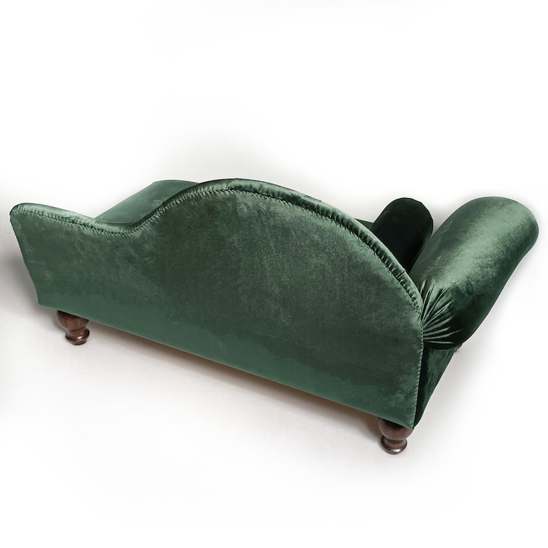 May include: A small, dark green velvet pet sofa featuring a curved backrest, a rolled armrest on one side, and dark brown wooden bun feet. The upholstery has a soft, plush texture, and the frame is shaped with a distinct, elegant silhouette.