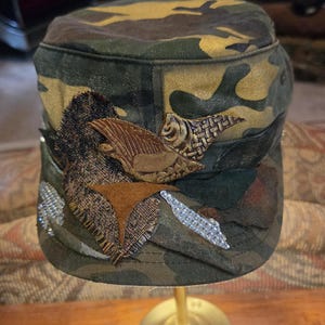 May include: Camouflage-patterned cap with decorative embellishments. The cap features a mix of green, brown, and tan hues. It is adorned with textured patches, including a brown leather-like eagle and sparkling rhinestones, adding a unique touch to the military-style hat.