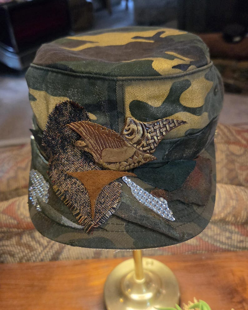 May include: Camouflage-patterned cap with decorative embellishments. The cap features a mix of green, brown, and tan hues. It is adorned with textured patches, including a brown leather-like eagle and sparkling rhinestones, adding a unique touch to the military-style hat.
