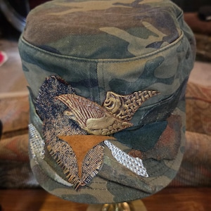 May include: Camouflage-patterned cap with decorative patches and a textured, bronze-colored emblem. The cap features a flat top and a curved brim, with various fabric embellishments in shades of green, brown, and tan. The emblem appears to be a stylized bird.