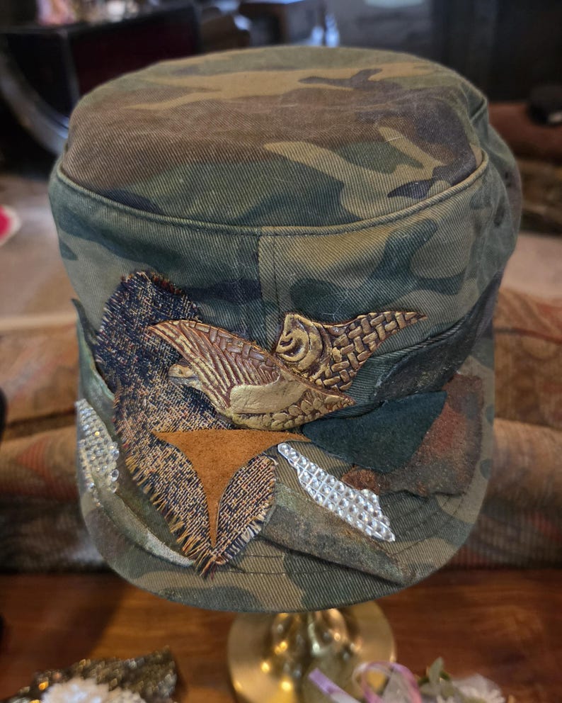 May include: Camouflage-patterned cap with decorative patches and a textured, bronze-colored emblem. The cap features a flat top and a curved brim, with various fabric embellishments in shades of green, brown, and tan. The emblem appears to be a stylized bird.
