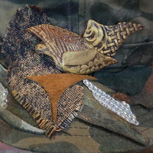 May include: Camouflage-patterned hat with a decorative bird and fabric embellishments. The bird is brown and gold, with textured details. Additional accents include a brown suede shape and a line of clear rhinestones.