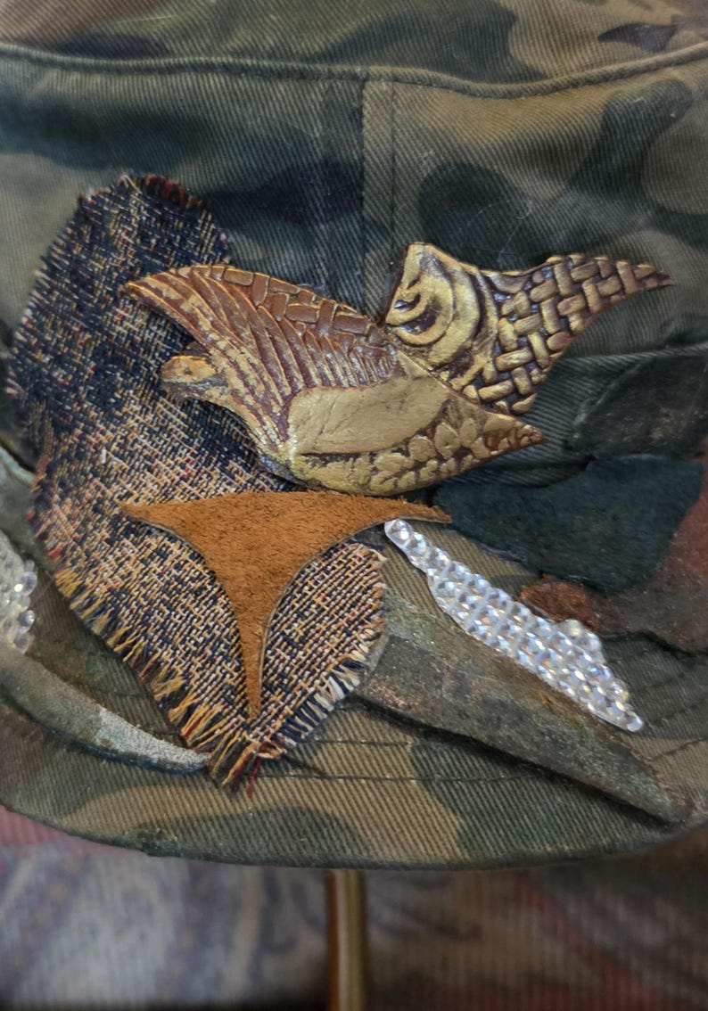May include: Camouflage-patterned hat with a decorative bird and fabric embellishments. The bird is brown and gold, with textured details. Additional accents include a brown suede shape and a line of clear rhinestones.