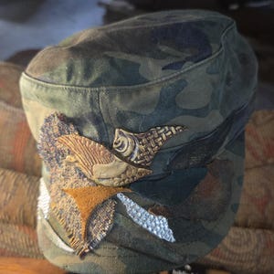 May include: Camouflage print military-style cap with decorative embellishments. The cap features a mix of textures, including leather and woven details, in shades of brown, tan, and silver. The cap's brim is slightly curved, and the overall design is a blend of utility and artistic flair.