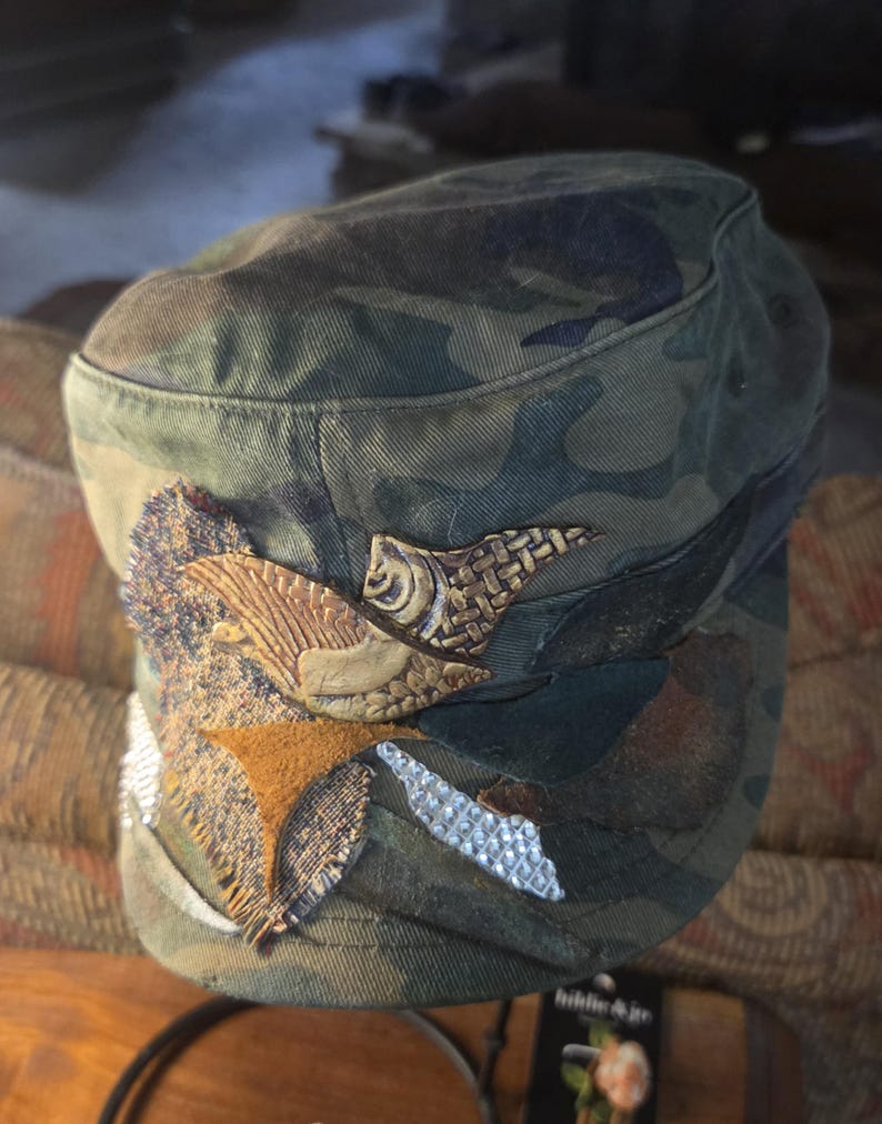 May include: Camouflage print military-style cap with decorative embellishments. The cap features a mix of textures, including leather and woven details, in shades of brown, tan, and silver. The cap's brim is slightly curved, and the overall design is a blend of utility and artistic flair.